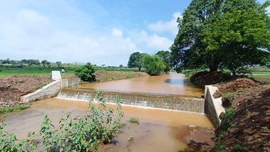 Small concrete irrigation weir channeling muddy river water through green farmland under a clear sky.