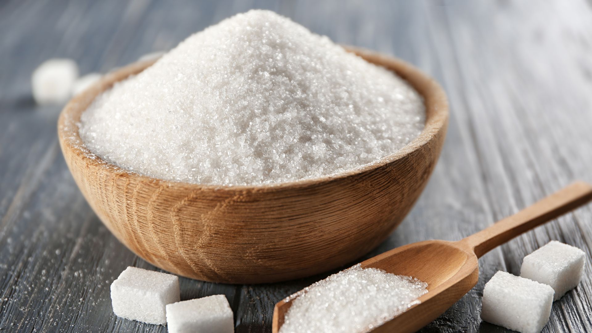 A wooden bowl heaped with sugar, next to a wooden scoop filled with sugar and sugar cubes.